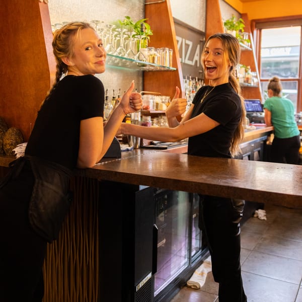 Two smiling Laziza baristas give thumbs up behind the bar, with glasses and bottles in the background.