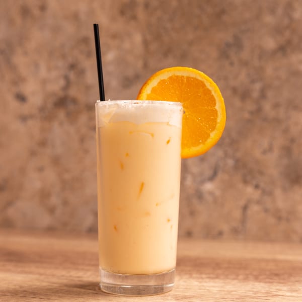A tall glass of creamy orange drink with ice, a black straw, and an orange slice garnish.