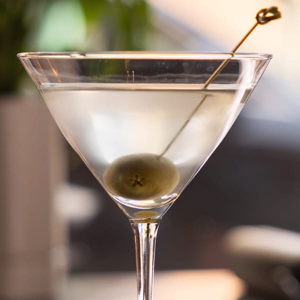 A martini in a clear glass with a green olive on a pick, against a blurred background.