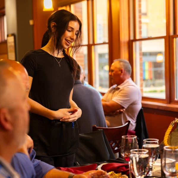 A smiling Laziza server in a black uniform stands by a table with guests and drinks.