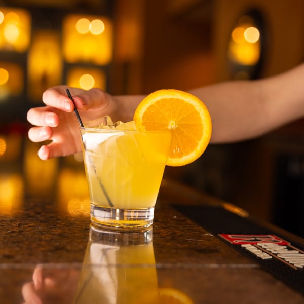 A hand stirs a yellow cocktail with ice and an orange slice on a bar counter.
