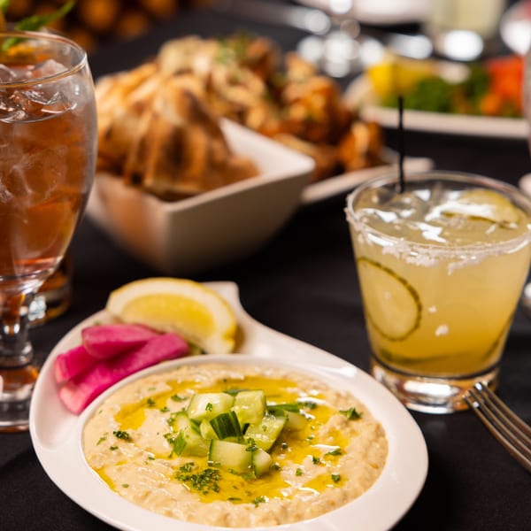 Laziza's hummus with cucumber, pickled turnips, lemon, and cocktails on a dark table.