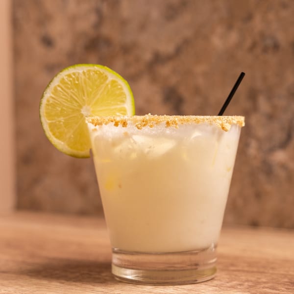 A creamy white cocktail with a graham cracker rim, lime slice, and black straw.