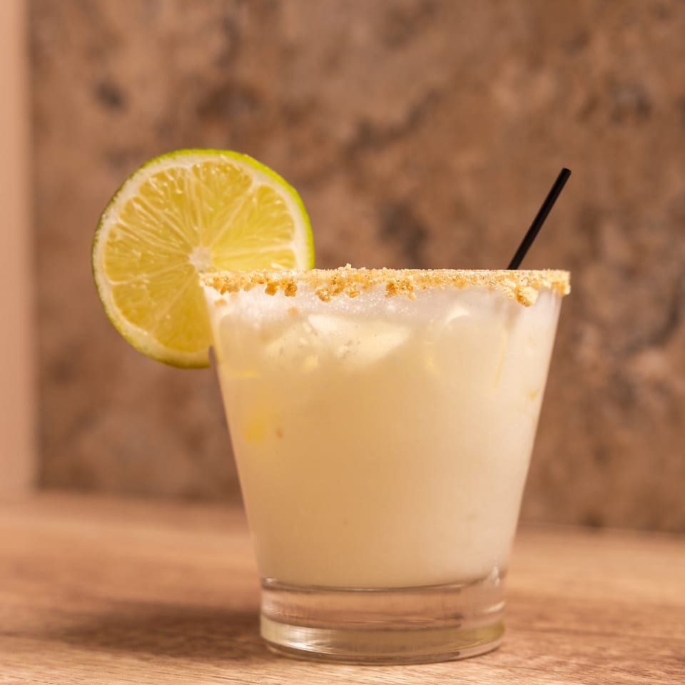 A creamy white cocktail with a graham cracker rim, lime slice, and black straw.