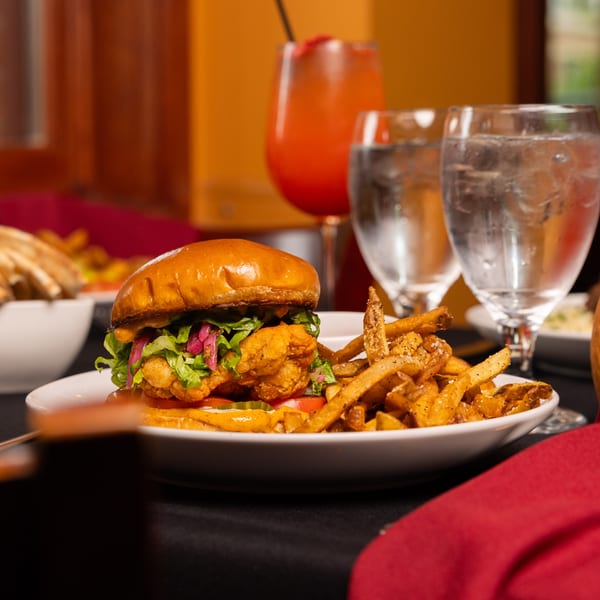 A Laziza fried chicken sandwich with fries, a red cocktail, and water glasses on a dark table.