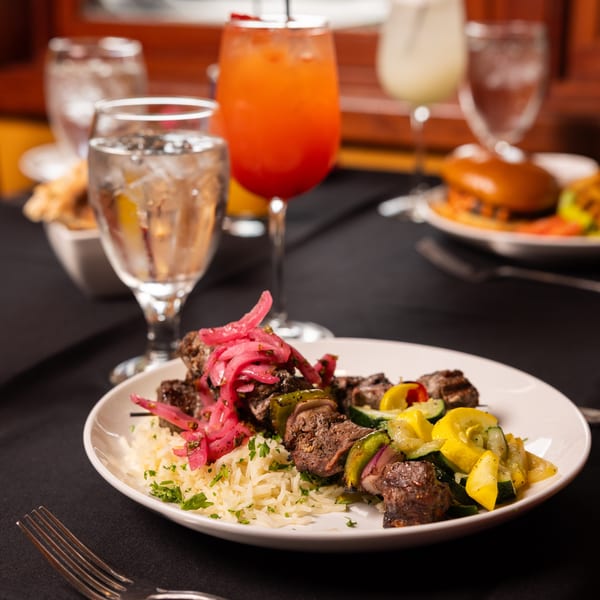 A Laziza beef skewer with rice, pickled onions, and grilled vegetables, with drinks in the background.