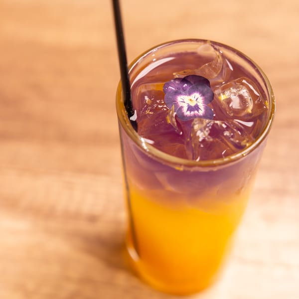 A vibrant two-toned drink with a purple edible flower and ice, served in a tall glass.