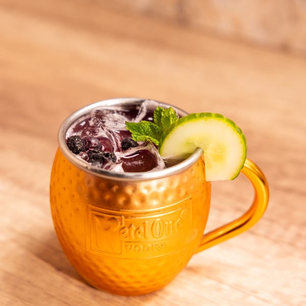A copper Moscow Mule mug filled with a dark beverage, ice, blueberries, mint, and a cucumber slice.
