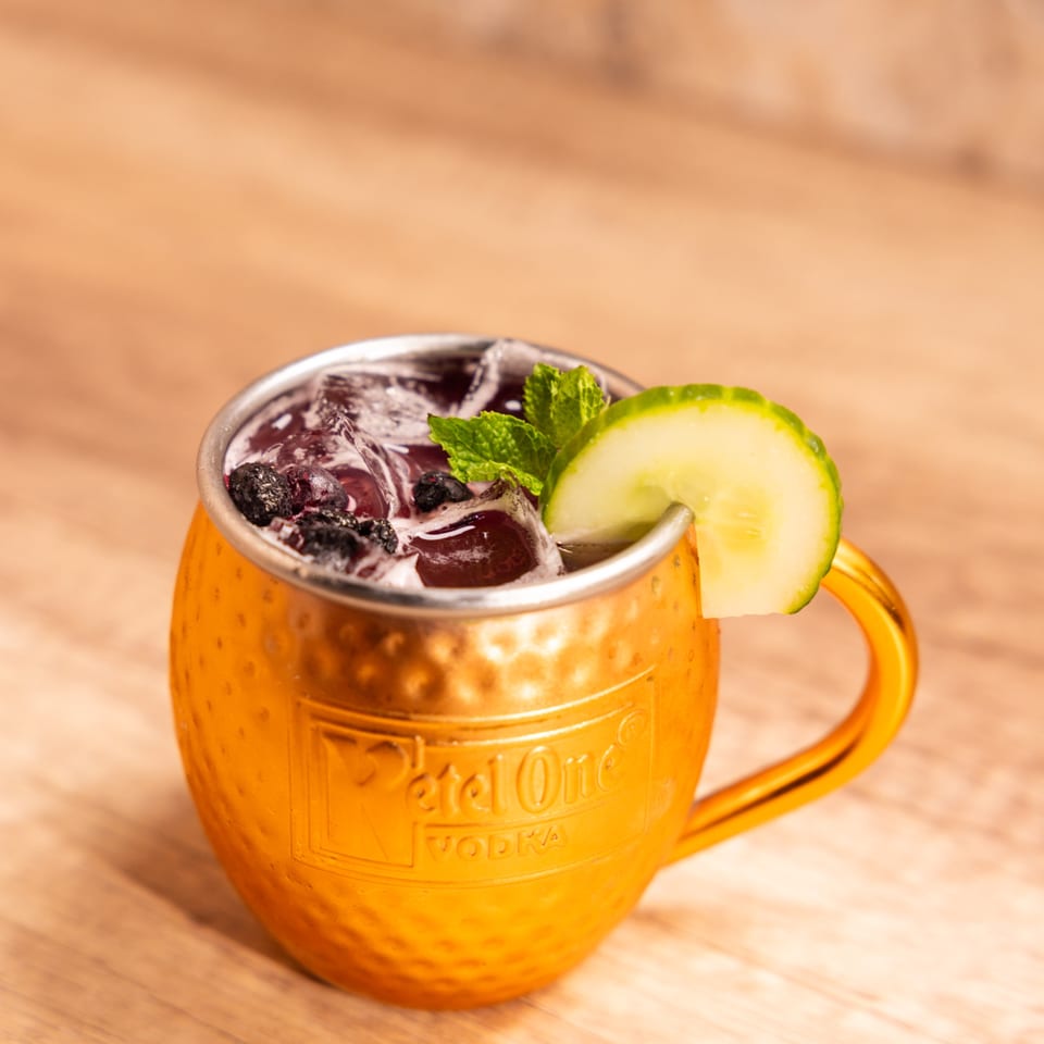 A copper Moscow Mule mug filled with a dark beverage, ice, blueberries, mint, and a cucumber slice.