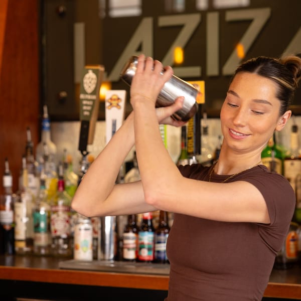 A smiling bartender at Laziza shakes a cocktail shaker behind a well-stocked bar.